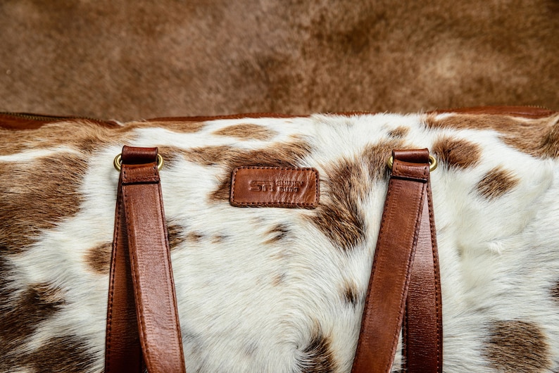 May include: A duffel bag featuring a white and brown cowhide pattern with smooth brown leather handles and a rectangular leather patch embossed with the text ST. SIMON BAG. The bag includes gold-toned metal hardware connecting the straps.