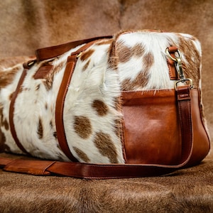 May include: A duffel bag featuring a white and brown cowhide pattern on the main body, accented with smooth brown leather side panels, handles, and a detachable shoulder strap with brass-toned metal hardware clips and buckles.