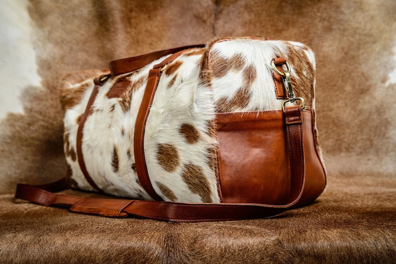 May include: A duffel bag featuring a white and brown cowhide pattern on the main body, accented with smooth brown leather side panels, handles, and a detachable shoulder strap with brass-toned metal hardware clips and buckles.