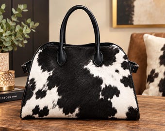 May include: A handbag featuring a black and white cowhide pattern with a textured fur-like finish. The bag has two black leather handles attached to the top, a curved upper silhouette, and side straps with metal hardware accents on both ends.