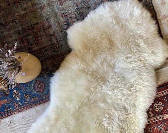 May include: A white sheepskin rug with a soft, fluffy texture. The rug is laid out on a patterned rug with red, blue, and white colors.