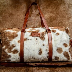 May include: A rectangular duffel bag featuring a white and brown cowhide pattern with a textured fur finish. The bag includes smooth brown leather handles and a matching adjustable shoulder strap, with a small rectangular leather brand patch.