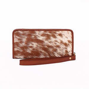 May include: A brown leather wallet with a white and brown cowhide pattern. The wallet has a zipper closure and a wrist strap.