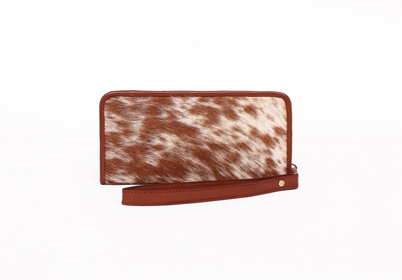 May include: A brown leather wallet with a white and brown cowhide pattern. The wallet has a zipper closure and a wrist strap.