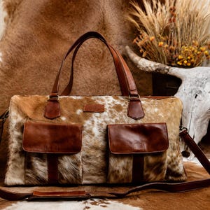 May include: A duffel bag crafted from brown and white cowhide with smooth brown leather accents. The bag features two front pockets with flap closures, two top handles, and a detachable shoulder strap with a padded leather shoulder rest.