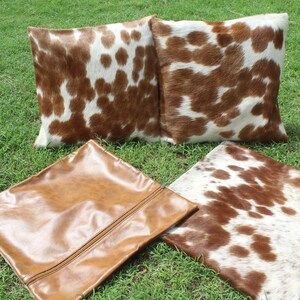 May include: Four decorative throw pillows with a cowhide pattern. Two pillows are brown and white, one is brown leather, and one is white with brown spots.