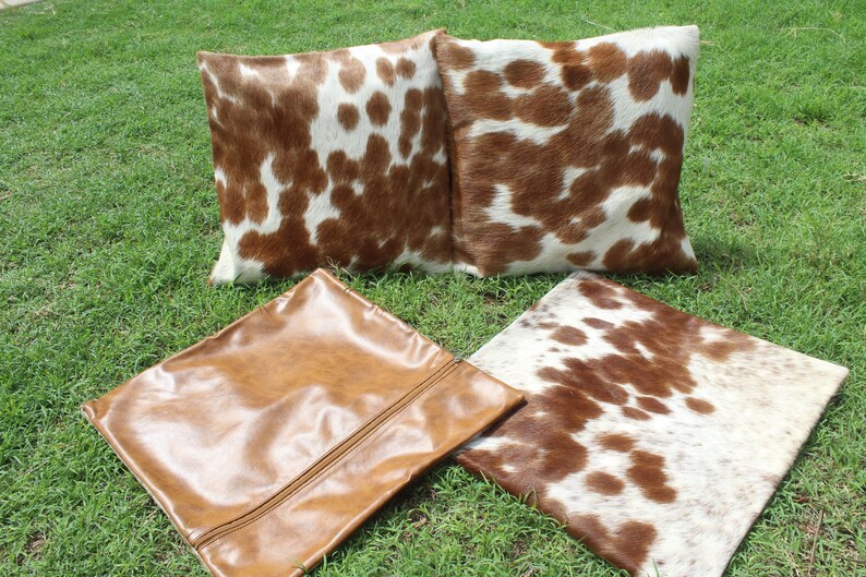 May include: Four decorative throw pillows with a cowhide pattern. Two pillows are brown and white, one is brown leather, and one is white with brown spots.