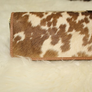 Puede incluir: Una cartera de piel de vaca marr&oacute;n y blanca con un tirador de cremallera de cuero marr&oacute;n.