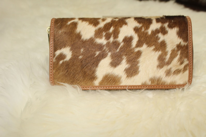 Puede incluir: Una cartera de piel de vaca marr&oacute;n y blanca con un tirador de cremallera de cuero marr&oacute;n.