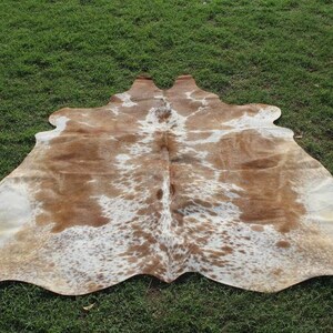 May include: A brown and white cowhide rug with a natural pattern. The rug is laid out on a green grassy surface.