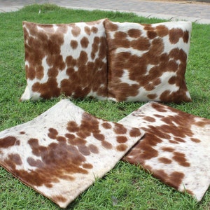 May include: Four brown and white cowhide throw pillows with a spotted pattern. The pillows are arranged on a green grassy surface.