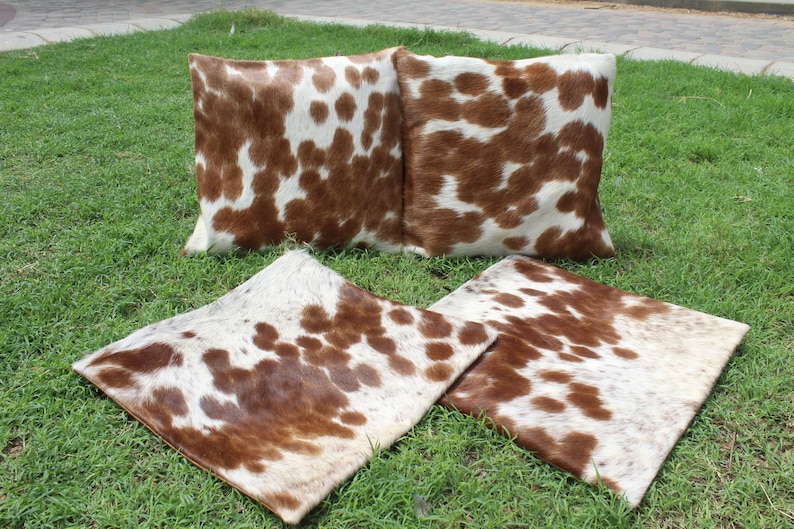 May include: Four brown and white cowhide throw pillows with a spotted pattern. The pillows are arranged on a green grassy surface.
