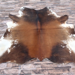 May include: A brown and white cowhide rug with a natural, spotted pattern. The rug is laid out on a stone floor.