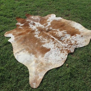 May include: A brown and white cowhide rug with a natural, spotted pattern. The rug is laid out on a green grassy surface.