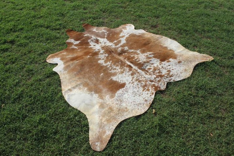 May include: A brown and white cowhide rug with a natural, spotted pattern. The rug is laid out on a green grassy surface.