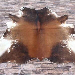 Op de afbeelding: Een bruin en wit koeienhuidkleed met een natuurlijk, gevlekt patroon. Het kleed ligt op een stenen vloer.
