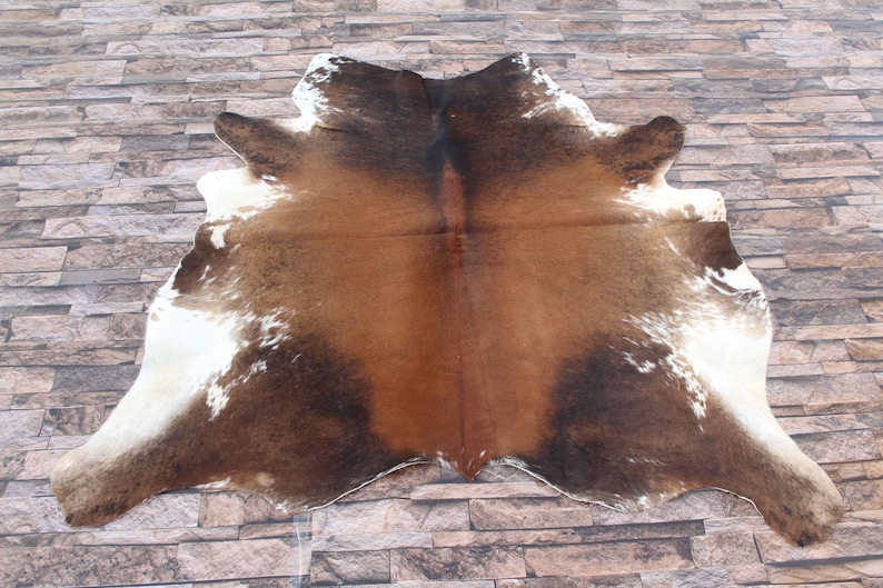 Op de afbeelding: Een bruin en wit koeienhuidkleed met een natuurlijk, gevlekt patroon. Het kleed ligt op een stenen vloer.