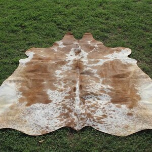 May include: A brown and white cowhide rug with a natural, spotted pattern. The rug is laid out on a green grassy surface.