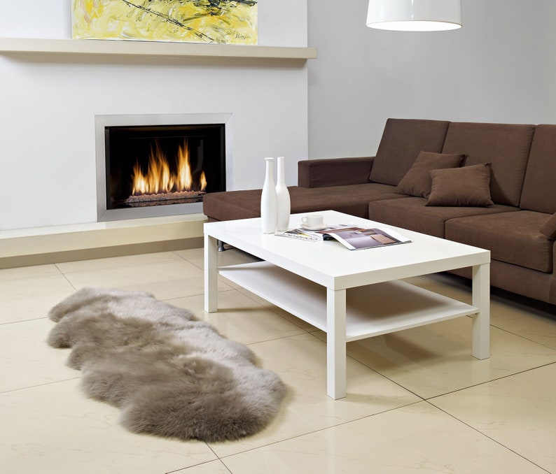 May include: A white coffee table with a lower shelf sits in a living room with a brown sectional sofa, a fireplace, and a beige sheepskin rug.
