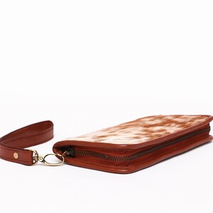 May include: A brown leather wallet with a white and brown cowhide pattern. The wallet has a zipper closure and a wrist strap.