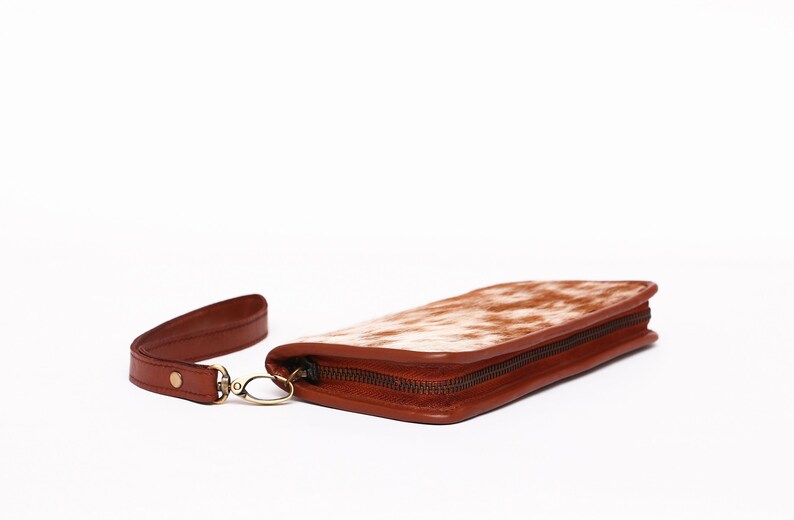 May include: A brown leather wallet with a white and brown cowhide pattern. The wallet has a zipper closure and a wrist strap.