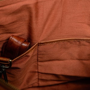 May include: The interior of a bag featuring a rust-colored fabric lining with a flat open pocket and a leather strap attached to a brass-toned metal swivel clasp. The lining has a visible texture and the pocket is finished with a leather trim.