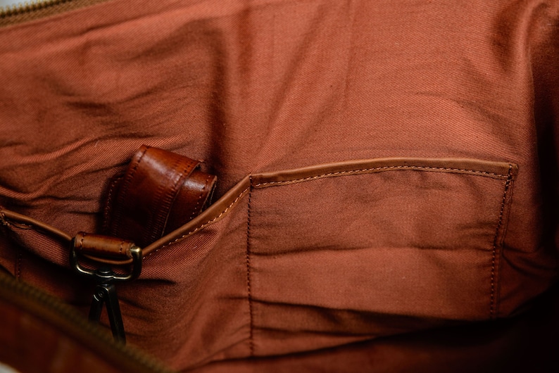 May include: The interior of a bag featuring a rust-colored fabric lining with a flat open pocket and a leather strap attached to a brass-toned metal swivel clasp. The lining has a visible texture and the pocket is finished with a leather trim.