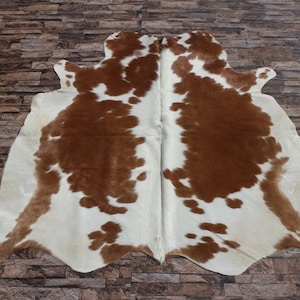 May include: A brown and white cowhide rug with a natural, spotted pattern. The rug is laid out on a stone floor.