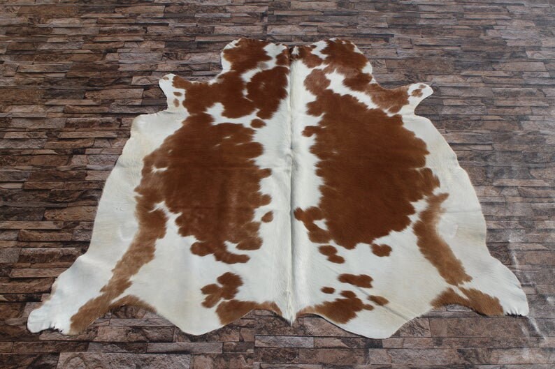 May include: A brown and white cowhide rug with a natural, spotted pattern. The rug is laid out on a stone floor.