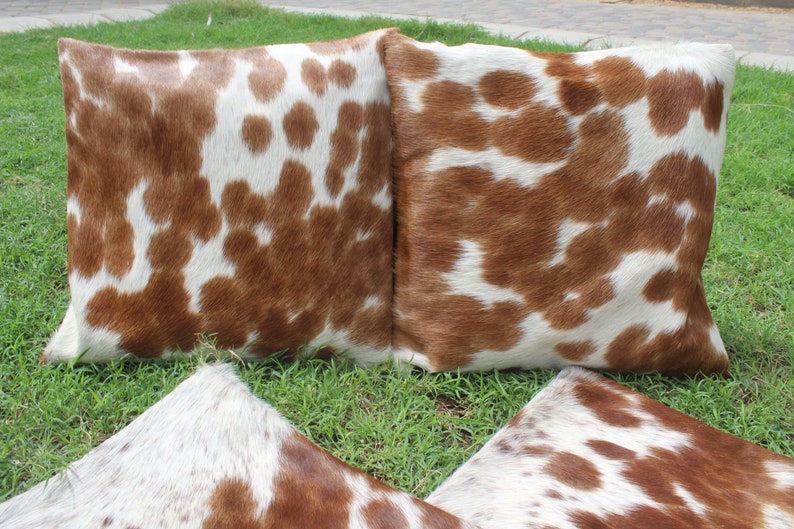 May include: Three brown and white cowhide throw pillows with a soft, fuzzy texture. The pillows are arranged in a triangular shape on a bed of green grass.