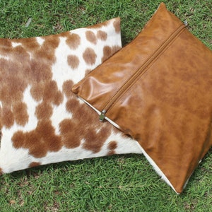 May include: Two decorative throw pillows. One pillow is brown faux leather with a zipper closure. The other pillow is made of brown and white cowhide.