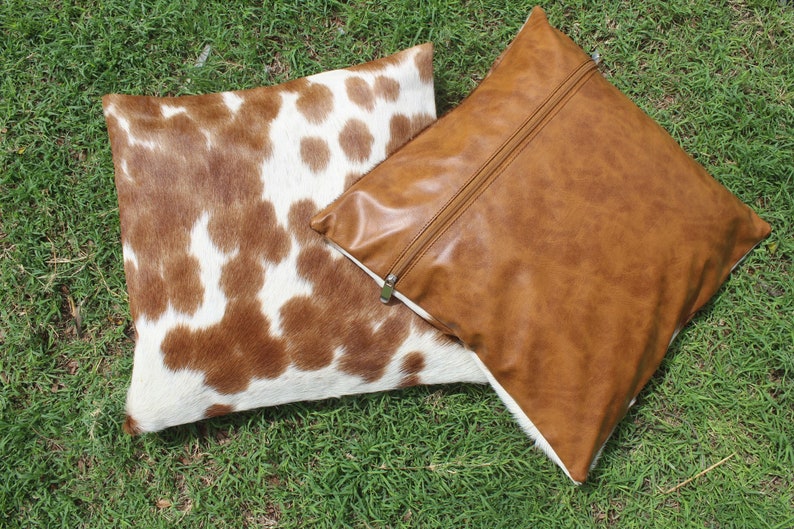 May include: Two decorative throw pillows. One pillow is brown faux leather with a zipper closure. The other pillow is made of brown and white cowhide.