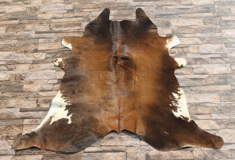 May include: A brown and white cowhide rug with a natural, rustic look. The hide has a variety of brown shades and white markings.