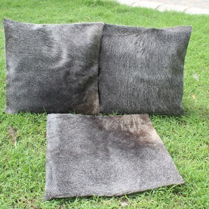 May include: Three gray cowhide throw pillows with a soft, fuzzy texture. The pillows are arranged on a green grassy surface.