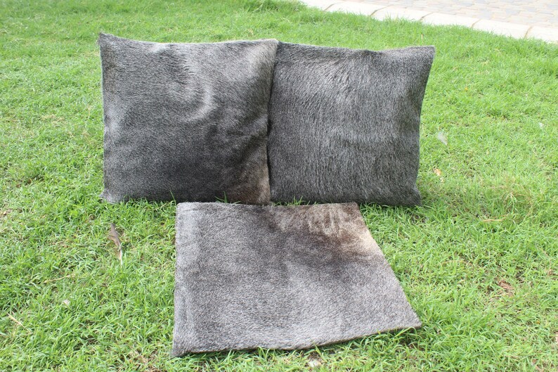 May include: Three gray cowhide throw pillows with a soft, fuzzy texture. The pillows are arranged on a green grassy surface.