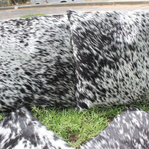 May include: Four black and white cowhide throw pillows with a soft, fuzzy texture. The pillows are arranged on a bed of green grass.