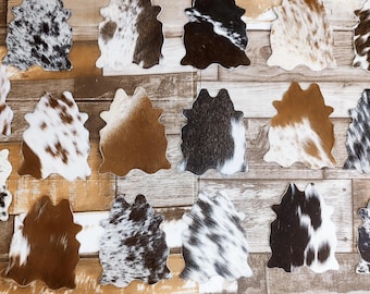 May include: A collection of cowhide coasters in various patterns and colors, including black and white speckled, brown and white, and solid brown. The coasters are arranged on a wooden background.