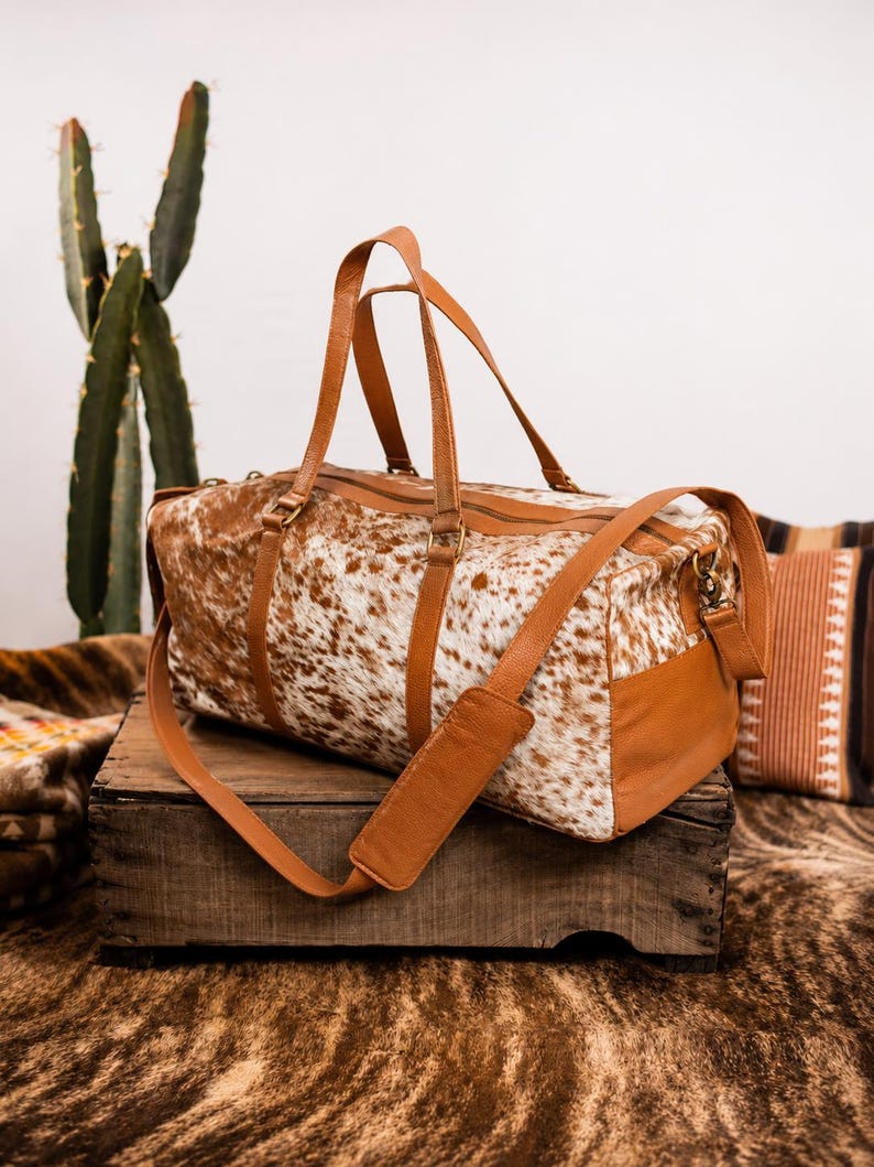 May include: A duffel bag featuring a white and brown cowhide pattern on the main body, complemented by tan leather handles, side panels, and an adjustable shoulder strap with a padded section. The bag includes a top zipper closure and metal hardware.