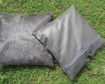 May include: Two square throw pillows. One pillow features a textured, dark gray, hair-on-hide surface. The second pillow is made of a smooth, solid gray synthetic material and includes a visible gray zipper closure running diagonally across the front.