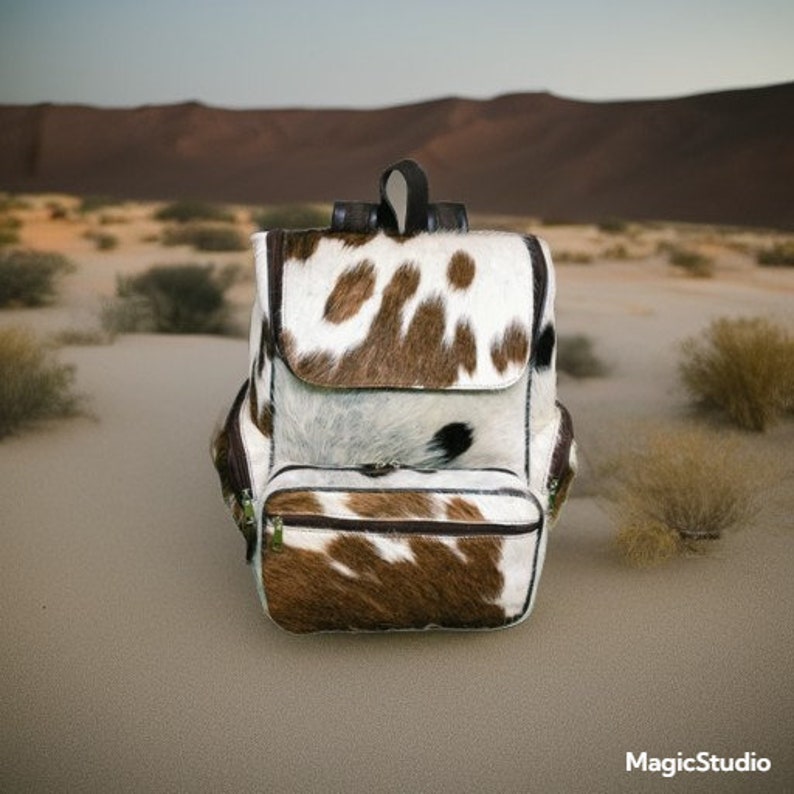 May include: A brown and white cowhide backpack with a brown leather trim. The backpack has a large main compartment and a smaller front pocket. The backpack is sitting on a sandy surface.