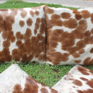 May include: Three brown and white cowhide throw pillows with a soft, fuzzy texture. The pillows are arranged in a triangular shape on a bed of green grass.