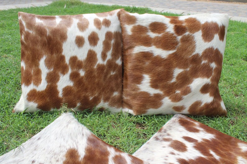 May include: Three brown and white cowhide throw pillows with a soft, fuzzy texture. The pillows are arranged in a triangular shape on a bed of green grass.