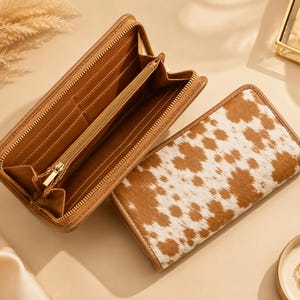 May include: A rectangular wallet with a brown and white cow print exterior and a tan interior. The wallet is shown in two positions: one is open to reveal multiple card slots and a zippered compartment, and the other is closed to show the patterned exterior.
