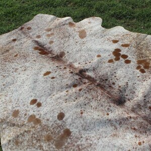 May include: A white cowhide rug with brown spots and a darker brown patch in the center. The rug is laid out on a green grassy surface.