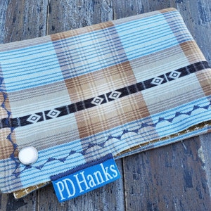 May include: A blue and brown plaid fabric wallet with a black and white geometric design and a silver button. The wallet has a blue label with the text "PD Hanks" on it.