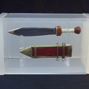 May include: A miniature Roman gladius sword with a wooden handle and a red leather scabbard. The sword and scabbard are displayed in a clear acrylic case.