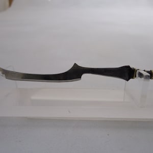 Puede incluir: Una pequeña espada de metal negro curvada con un mango dorado. La espada se muestra en un soporte de acrílico transparente.