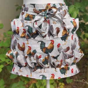 May include: A white apron with a rooster print and multiple pockets. The apron has a ruffled bottom and a bow tie.