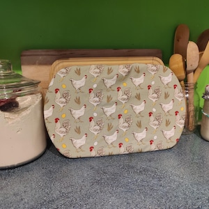 May include: A green and white fabric bread cover with a pattern of chickens and eggs. The cover is on top of a wooden breadboard.