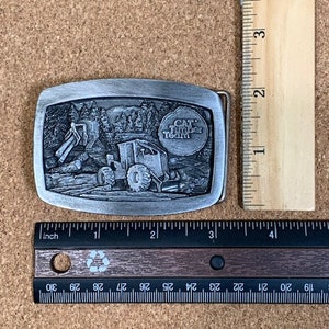 May include: A silver belt buckle featuring a detailed image of a construction vehicle and the text "CAT. THE BIG TEAM".